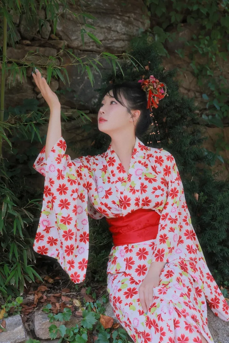 身着红花和服的女子坐于绿植石墙前，抬手轻触竹叶，气质清新温婉。