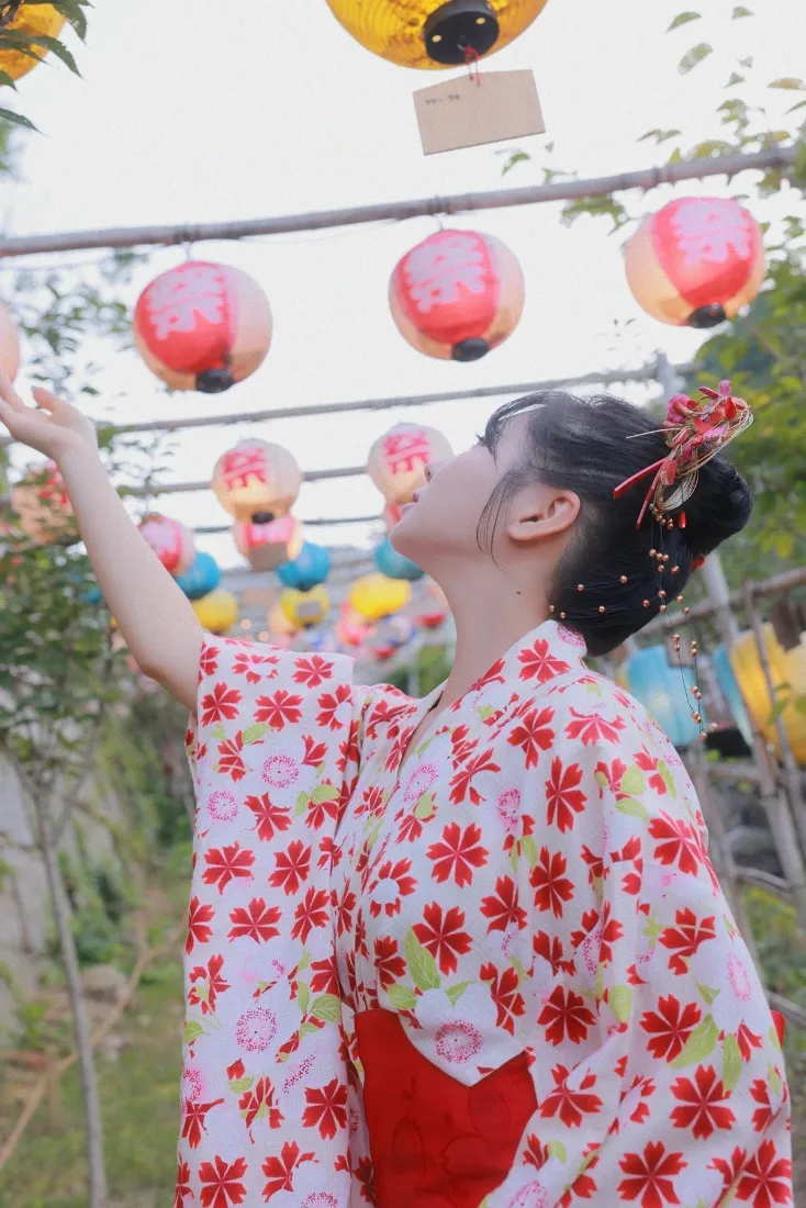 穿红花和服的女子在户外彩色灯笼下抬手仰望，背景绿植虚化。