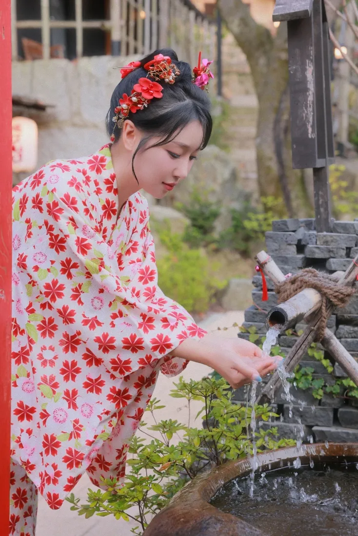 身着红樱花纹和服的女子在日式庭院竹制手水舍下俯身接水洗手，背景绿植与传统建筑虚化。