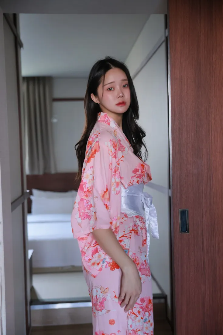 身着粉色印花和服的长发女子站在日式房间门口，背景可见床铺与窗帘