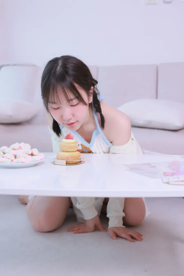 双马尾女孩跪于居家地板，俯身凑近桌上草莓小蛋糕，氛围温馨可爱。