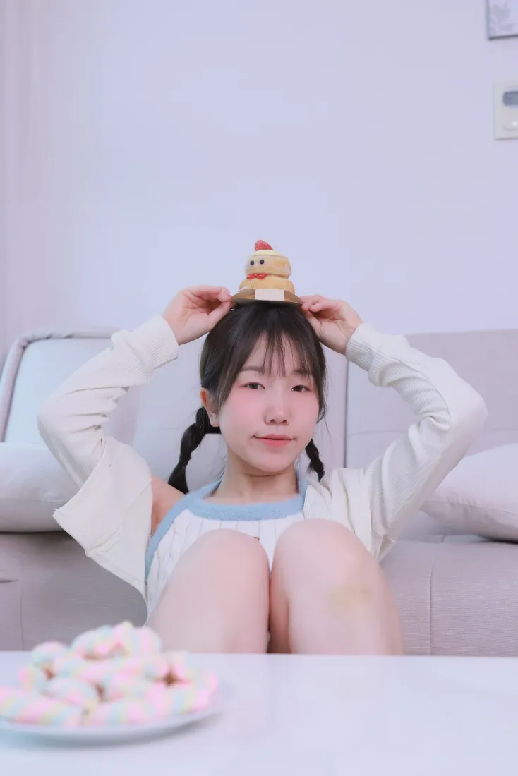双马尾女孩坐在居家沙发上，双手将小蛋糕摆件放在头顶，氛围甜美可爱。