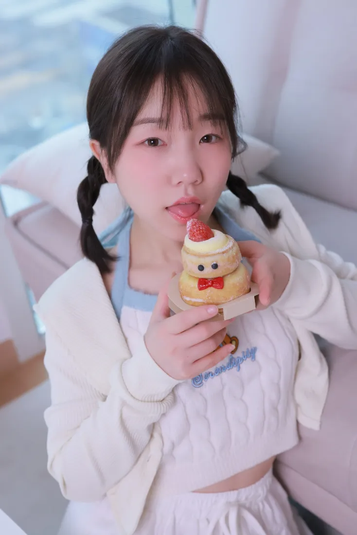 扎双麻花辫的白衣女孩坐在沙发上，手持卡通造型小蛋糕俏皮吐舌。