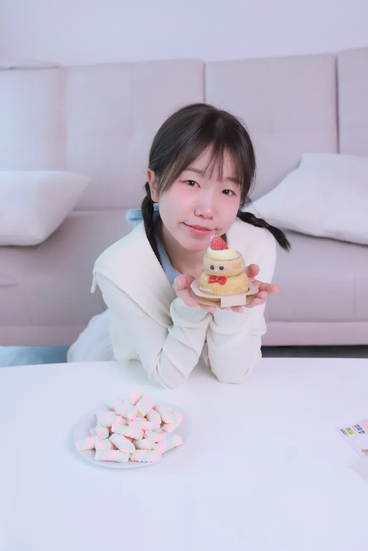 双马尾白衣女孩居家趴在白色桌面旁，双手捧着卡通造型小蛋糕。