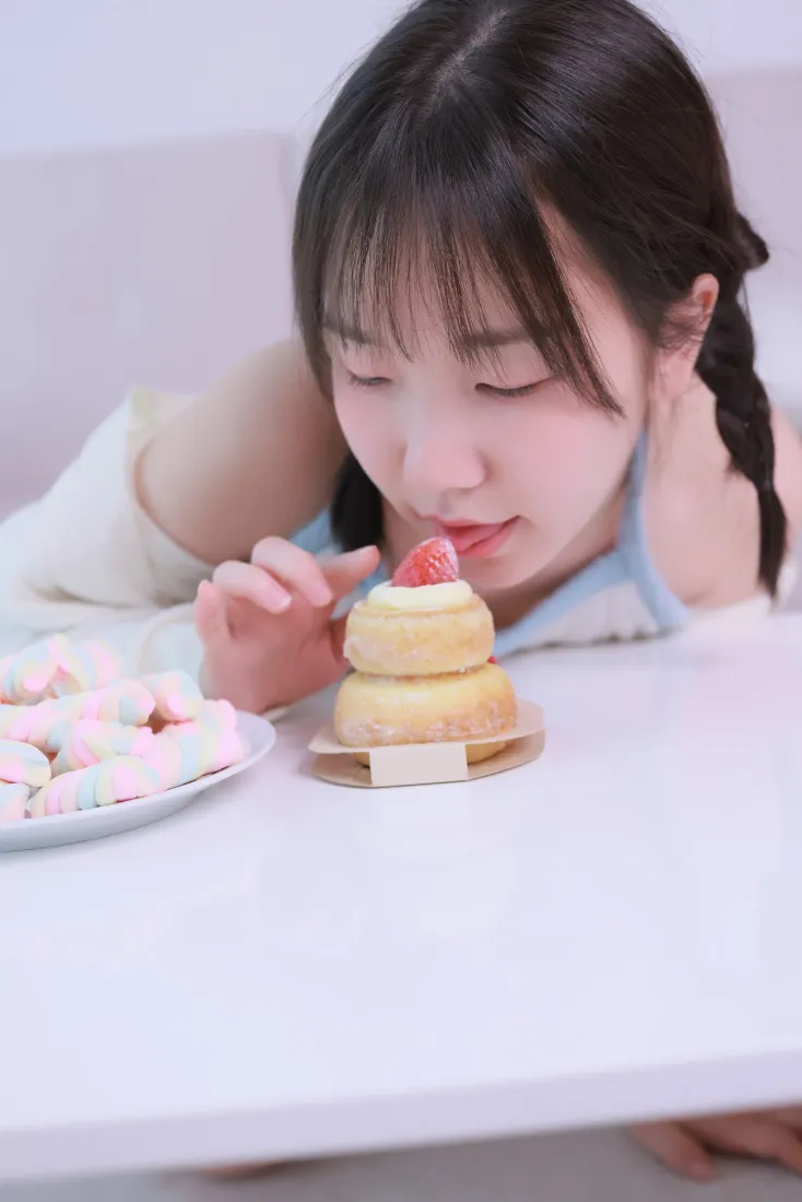 扎双麻花辫的少女趴在白色桌面旁，凑近草莓小蛋糕，旁侧摆放棉花糖