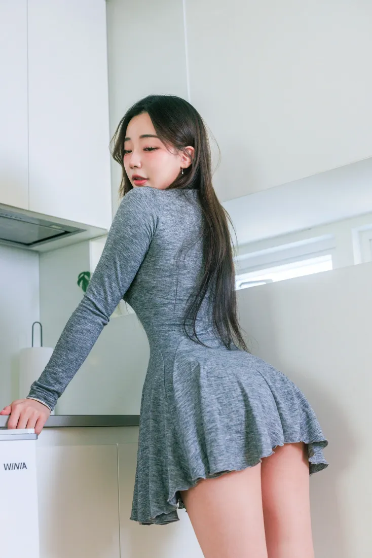 长发年轻女性身着灰色修身短裙，在明亮厨房内侧身回头倚靠台面。