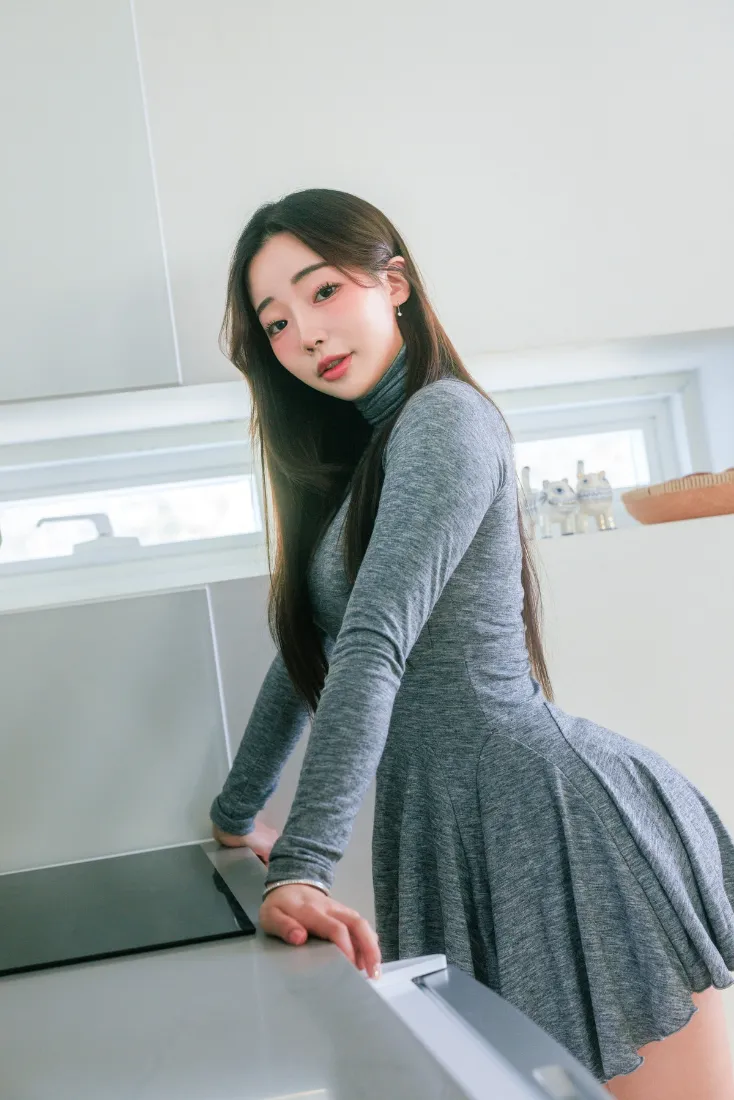 长发年轻女子身着灰色高领修身连衣裙，倚靠在明亮的居家厨房台边。