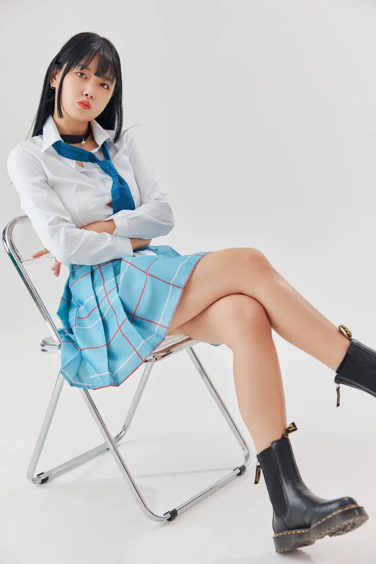 身着白衬衫蓝格裙校服的年轻女生，抱臂坐于金属折叠椅上，纯白简约背景。