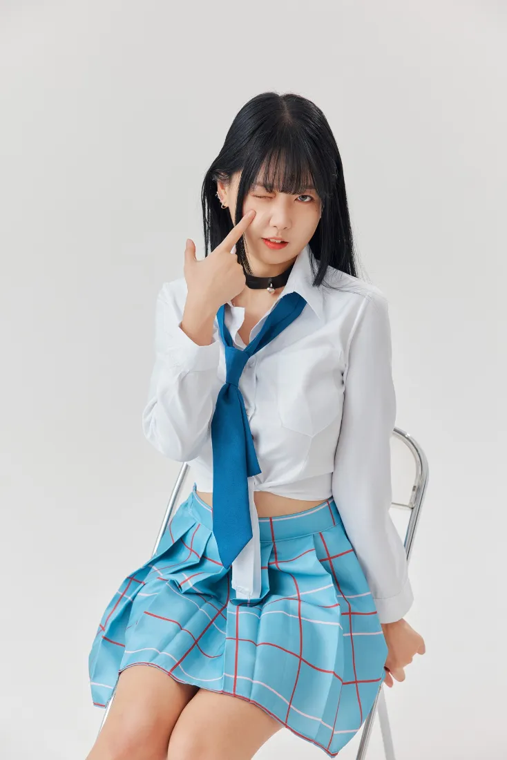 黑发少女穿白衬衫蓝格JK裙坐于椅上，眨眼比手势，浅灰纯色背景。