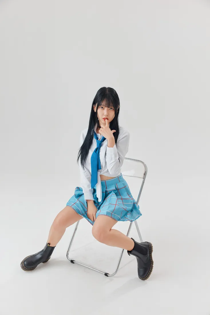 浅灰纯色背景下，黑长直少女着蓝白校服坐金属折叠椅，手指抵唇。