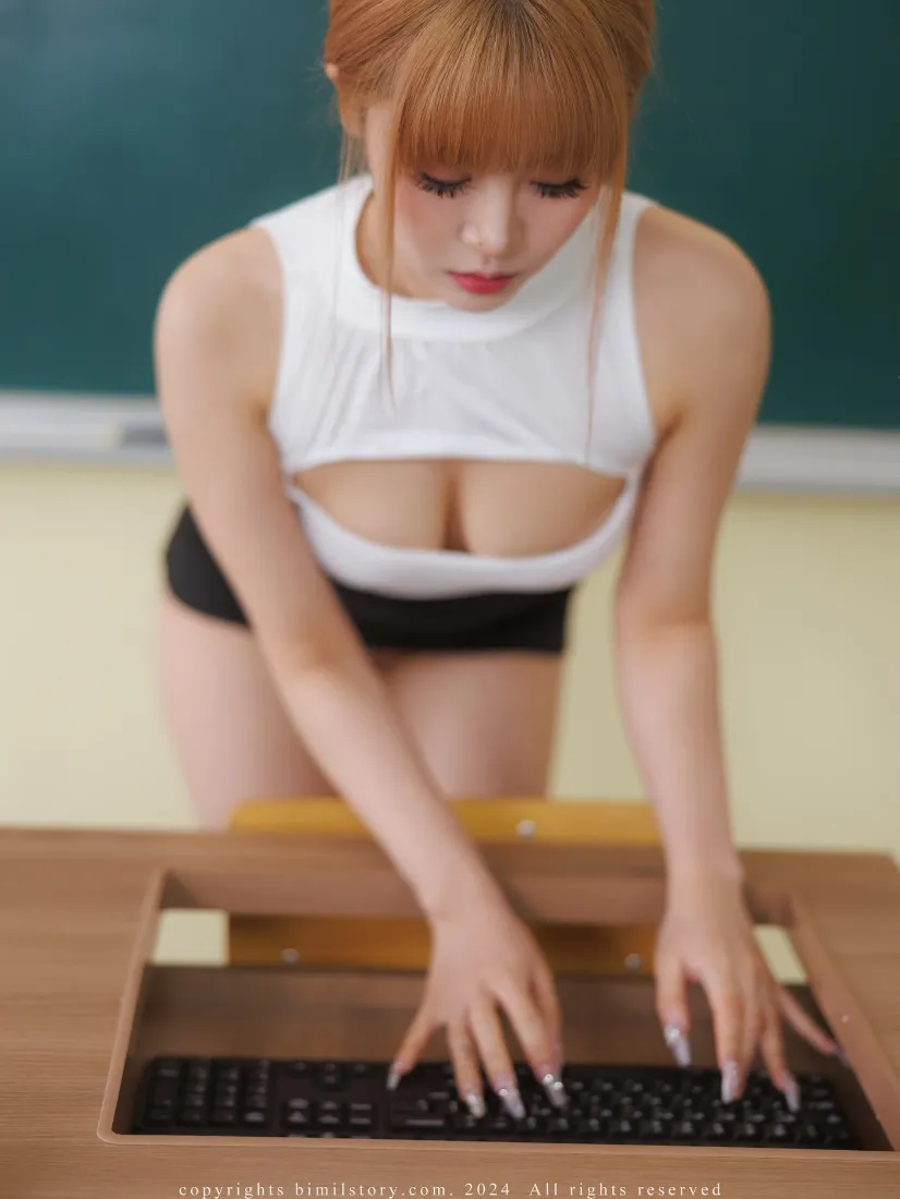 红发女性俯身于教室木质课桌前操作键盘，背景为绿色黑板