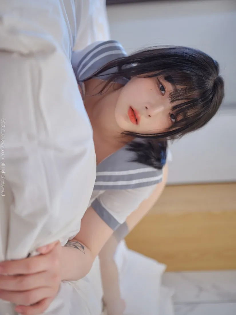 卧室床铺边，穿灰蓝水手服的短发红唇少女倚靠白色床品看向镜头