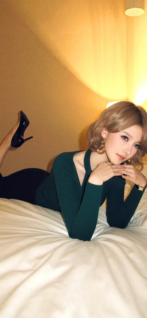 暖光卧室里，金发绿衣美女趴在白色床上，脚穿黑色高跟鞋。