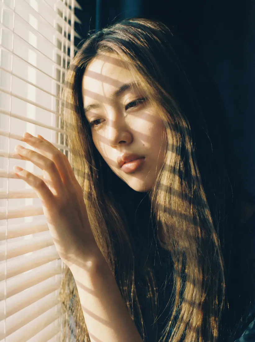 长发女生立于百叶窗边，暖光透过百叶窗在脸上投下条状光影。