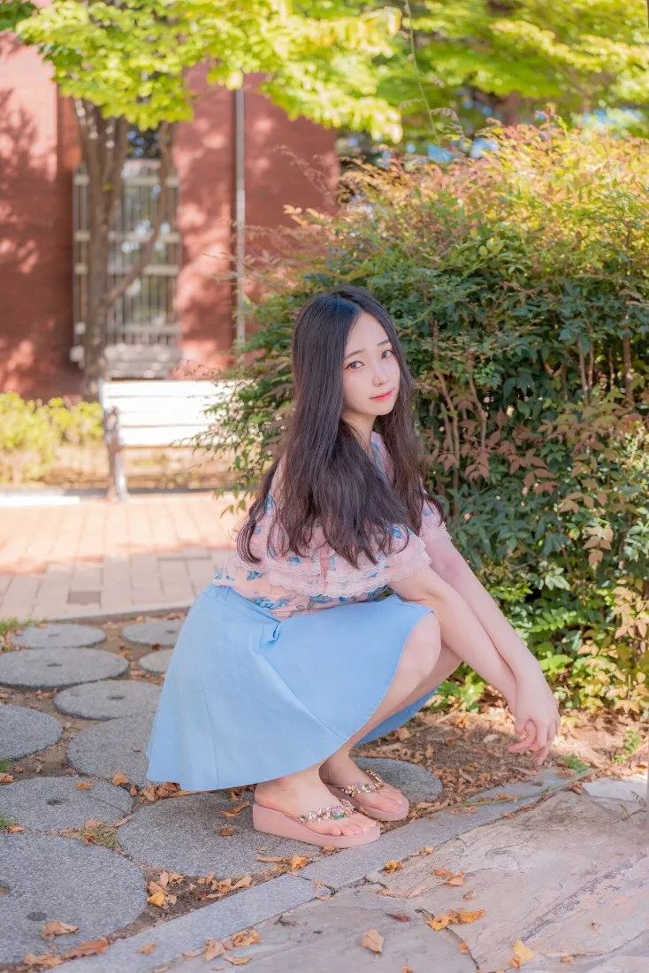 长发女生蹲在石板步道上，身着浅蓝裙，背景为绿植与红墙建筑。