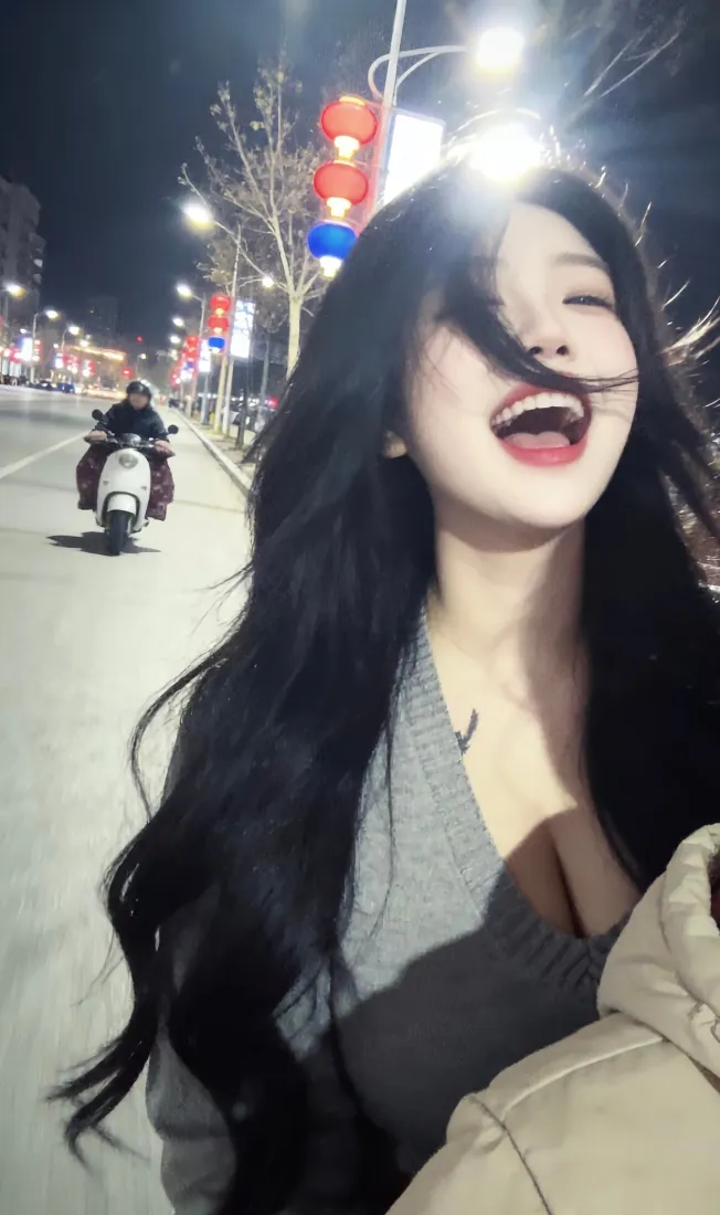 夜晚城市街道上，长发灰衣女子开心大笑，背景有路灯、红灯笼与电动车。