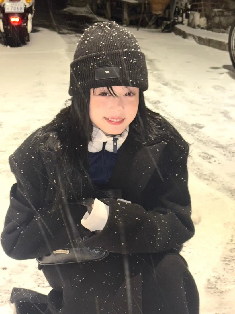 冬夜飘雪街头，戴针织帽穿黑大衣的黑发女生蹲坐微笑