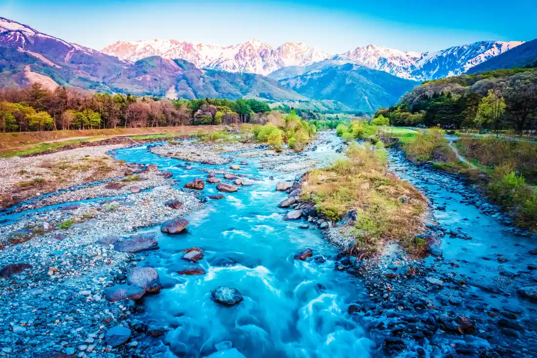 澄澈浅蓝溪流穿山谷而过，两岸林木葱郁，远处雪山连绵。