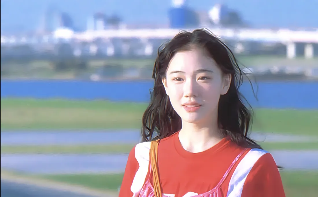 苍井优身着红衣站在河畔草地前，背景是蓝天与远处模糊的城市建筑。
