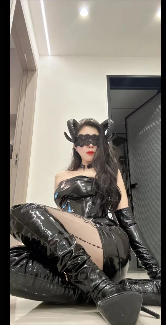 室内场景中，身着亮面黑色皮质套装、佩戴恶魔角与眼罩的魅魔cos女性席地而坐。