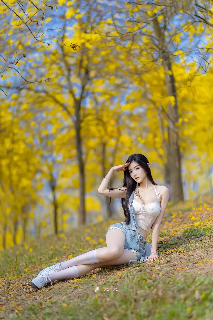 秋日黄叶林间，长发美女穿白吊带牛仔短裙坐于草坡上。