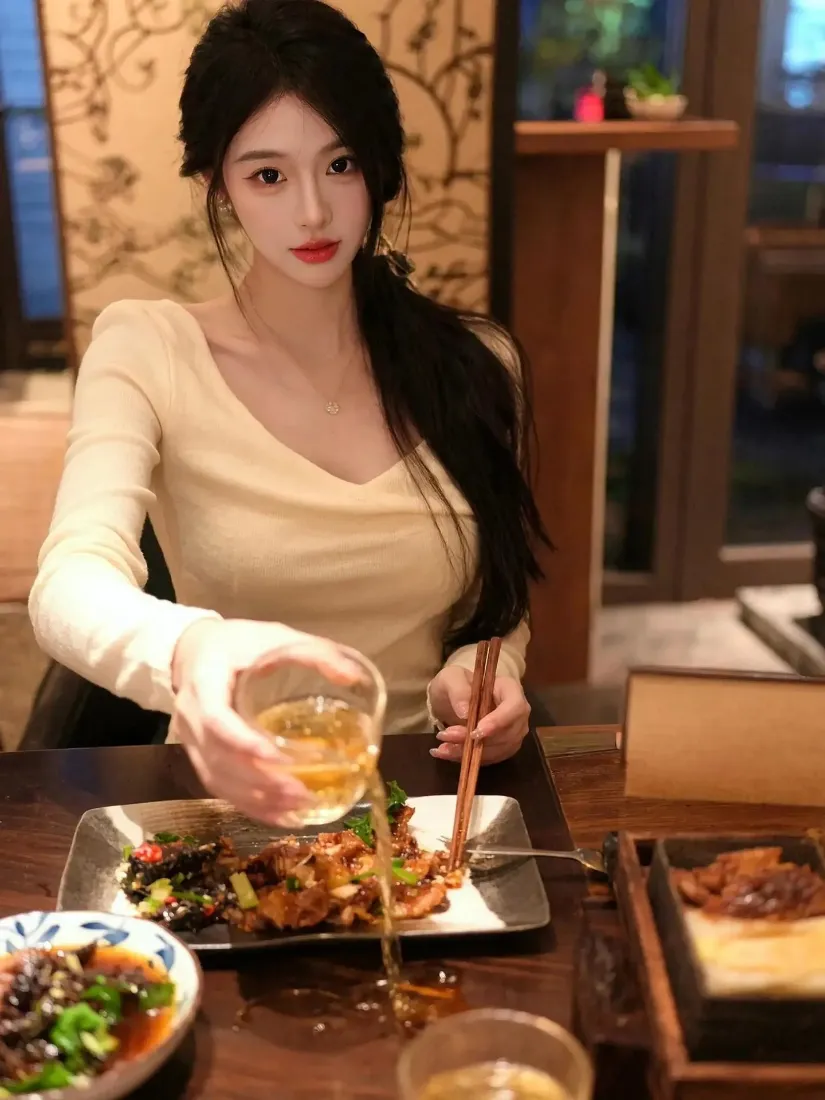 长发女子在暖光餐厅内，手持玻璃杯往餐盘倒饮品，桌上摆满中式菜肴