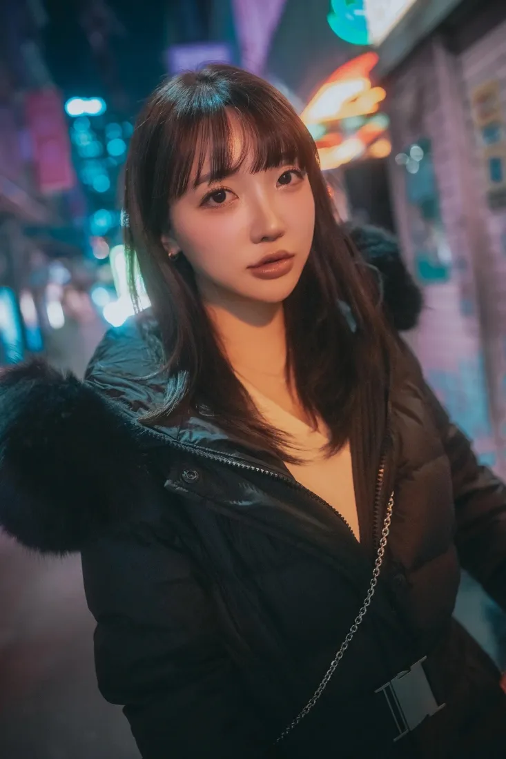 夜晚霓虹街道前，穿黑色毛领羽绒服的长发齐刘海女生半身街拍