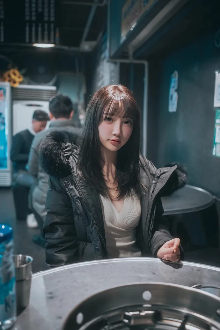 火锅店室内，穿黑色羽绒服的齐刘海长发女生坐在火锅桌前看向镜头。