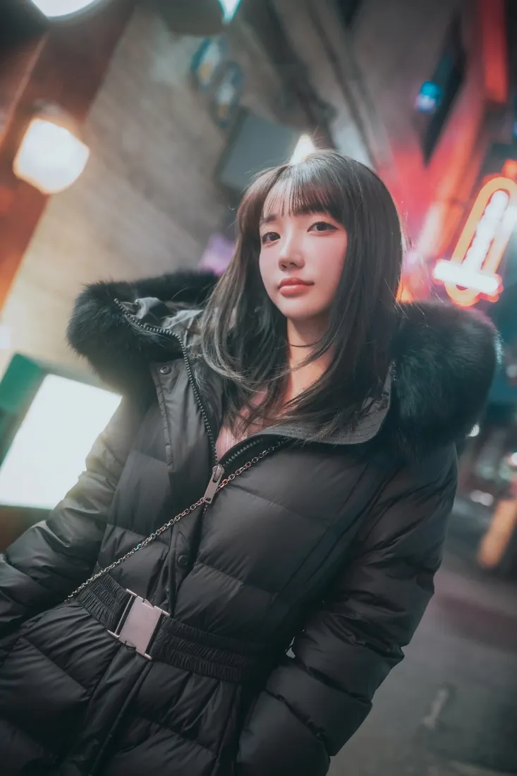冬日夜晚街头，齐刘海长发女生身着毛领黑色羽绒服，背景霓虹虚化。