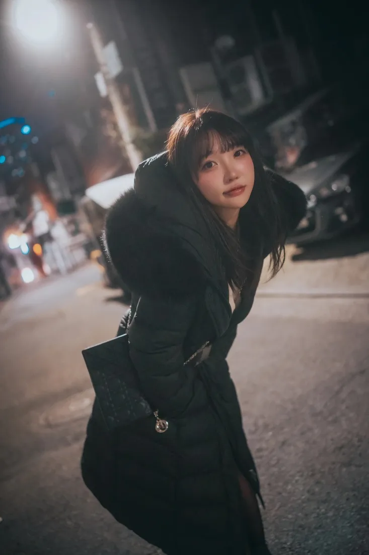 冬夜街头，穿黑色毛领大衣的年轻女子侧身回望，背景是虚化的街灯与街道。