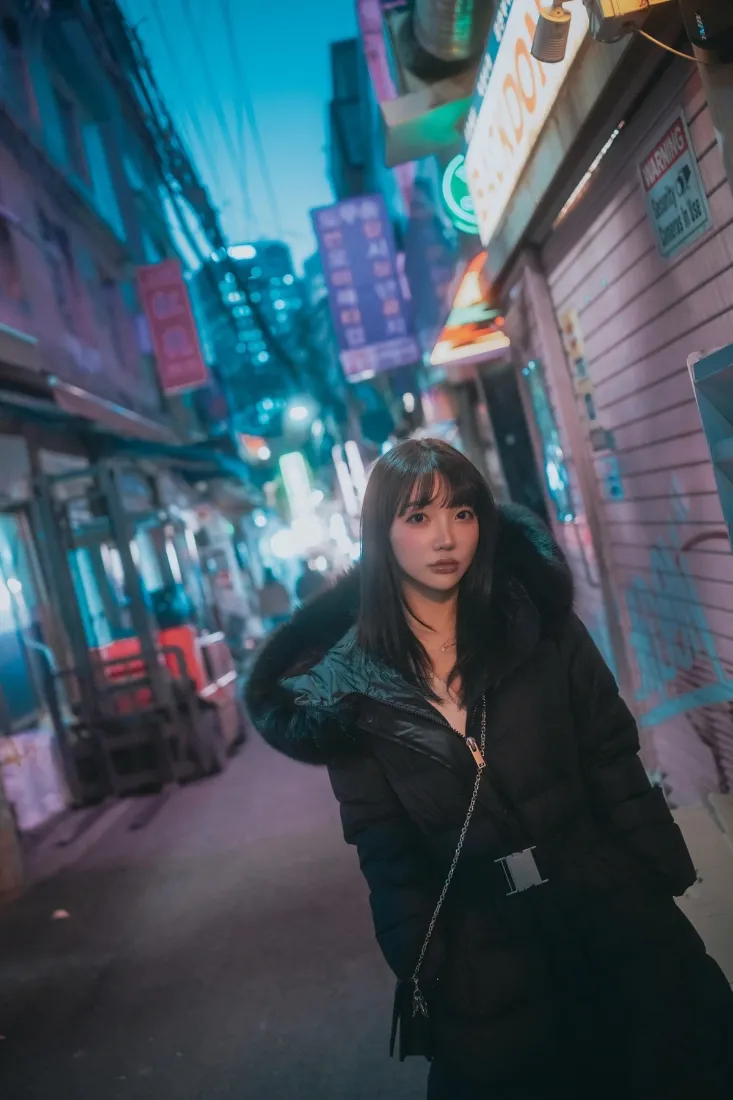 夜晚霓虹街巷中，穿黑色毛领皮衣的长发女子站在街道中央。