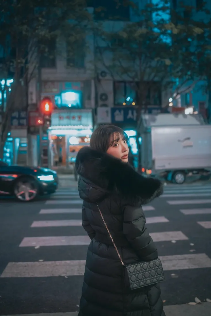夜晚城市街头，穿黑色毛领羽绒服的少女站在斑马线上回头，背景是霓虹街景与车辆。