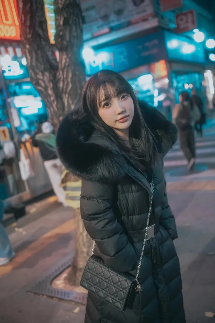 冬日夜晚霓虹街道上，穿毛领黑色羽绒服的年轻女生站在人行道上。
