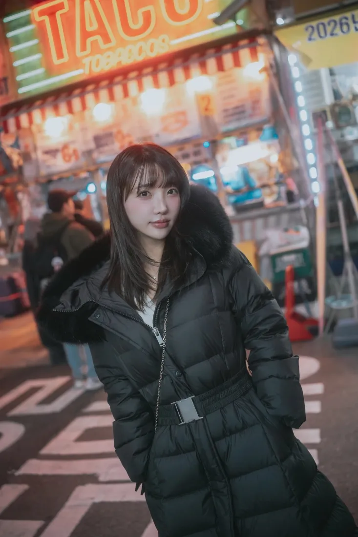 夜晚夜市街道上，穿黑色毛领羽绒服的长发年轻女子站在TACO招牌小吃摊前。