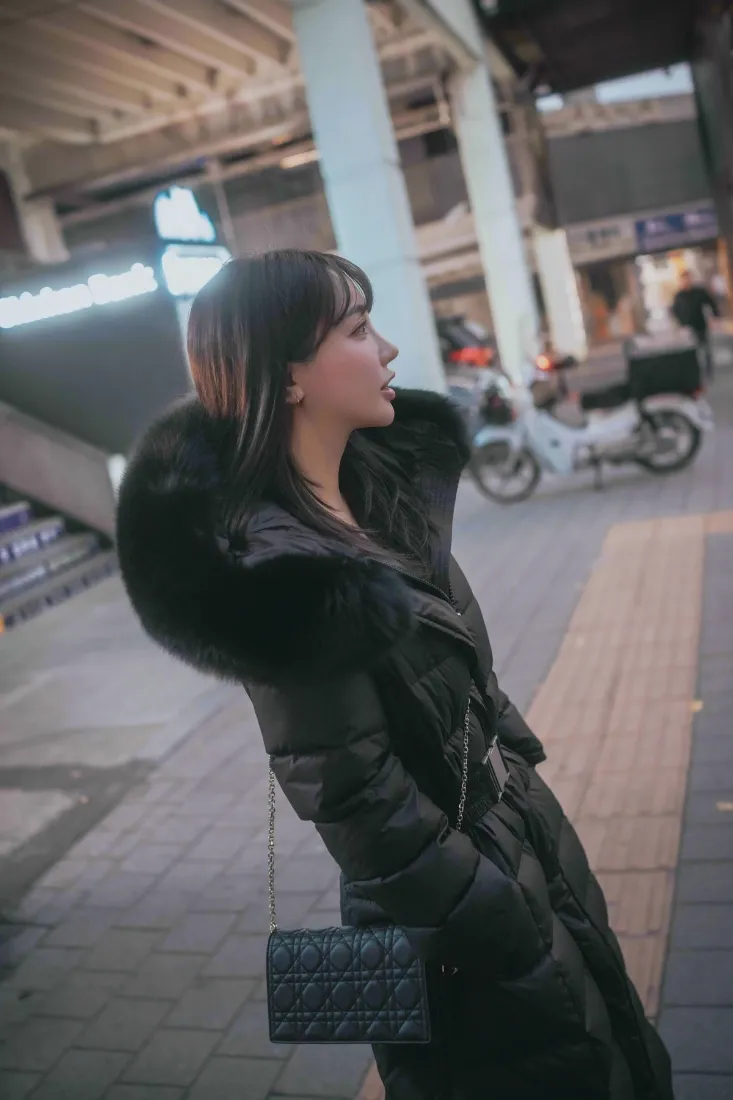 穿黑色毛领羽绒服的短发女生侧立在城市人行道上，背景虚化有电动车与建筑。