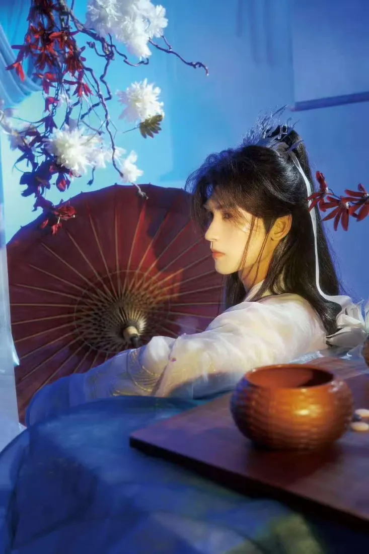 蓝调背景下白衣高马尾古装男子持红油纸伞，旁置陶罐与花枝。