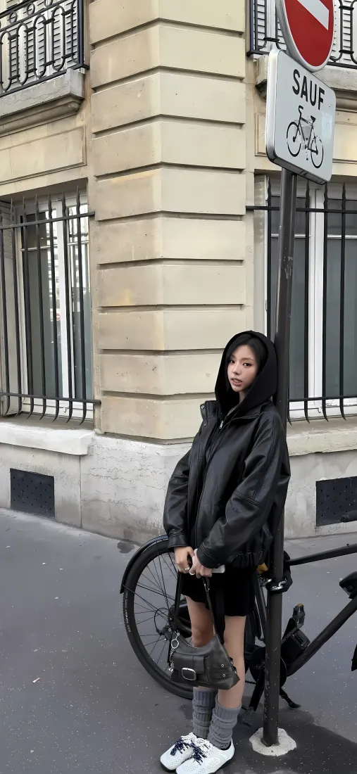黑衣女生靠在路牌杆旁，身旁停着自行车，背景是欧式街角建筑。