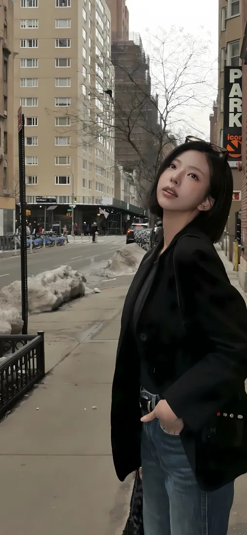 短发女子身着黑色外套牛仔裤，站在积雪的城市街道边回头望。