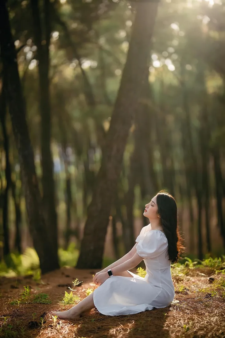 白裙少女坐于林间土地，仰头沐浴穿透林木的暖金色阳光。