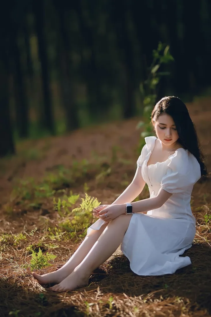 穿白裙的长发赤脚女子坐在林间暖光草地上，低头姿态静谧柔和。