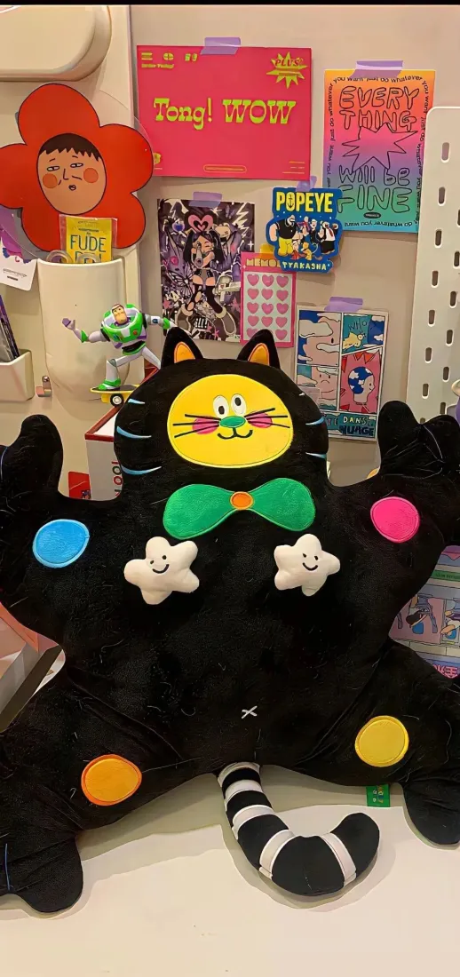 书桌前摆放着带彩色圆点、绿色领结的黑猫毛绒玩偶，背景墙面贴满装饰海报。