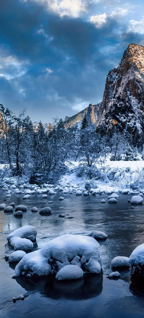 冬日河谷雪景，覆雪岩石静立河面，远处雪山与蓝天相映。