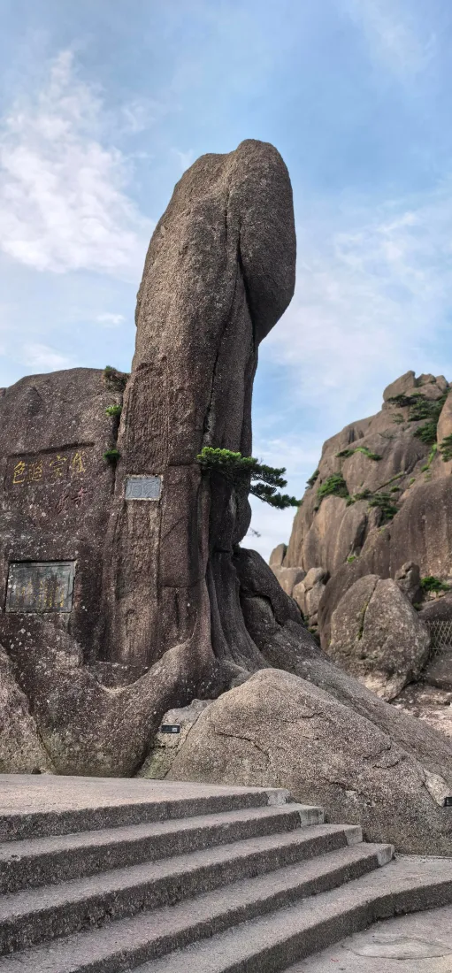 晴天山间，高耸的天然奇石矗立，旁有石阶与零星松树，背景是淡蓝天空。