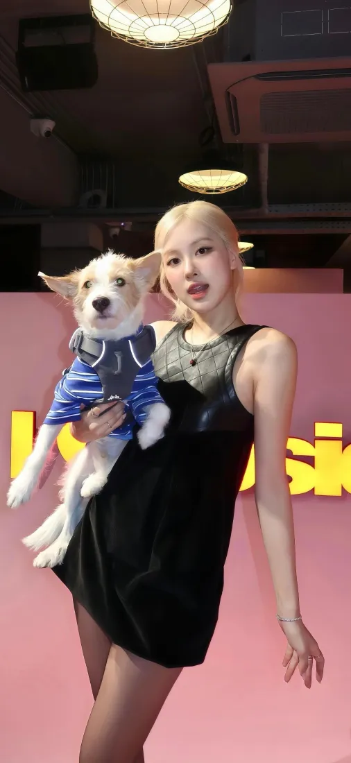 金发女星怀抱穿蓝条纹衣服的小狗，站在粉色背景墙前。