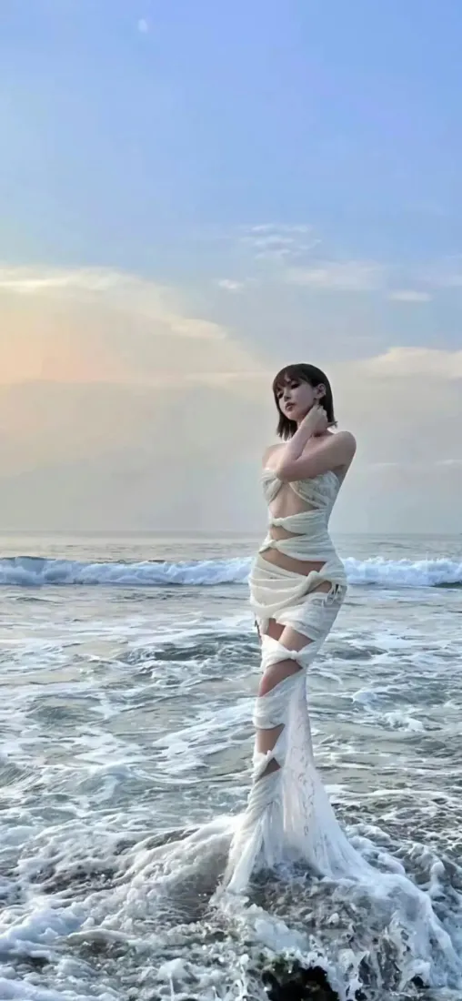 短发女子身着白色镂空纱裙立于黄昏浅海浪花中，背景是渐变蓝天与橙调天际。