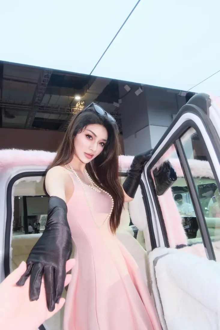 穿粉色无袖长裙戴黑长手套的长发美女站在打开车门的粉色豪车旁，伸手牵向镜头。