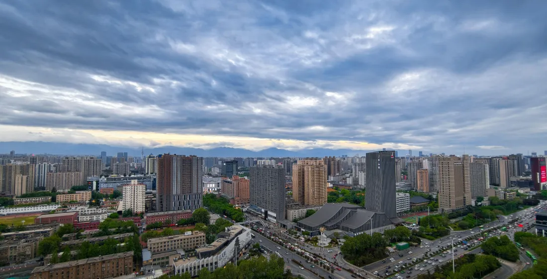 多云天空下的城市俯瞰景观，高楼林立道路纵横，远处可见朦胧山脉