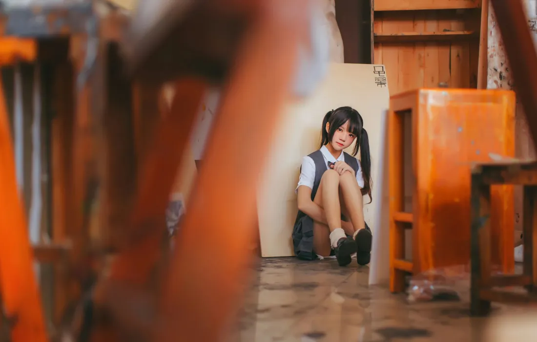 双马尾少女穿灰色JK校服抱膝坐在木质室内角落，前景虚化木椅