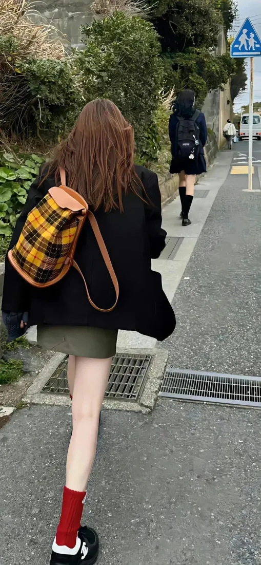 背黄格背包的长发女生背对镜头走在人行道上，身旁有绿植，前方还有行人。