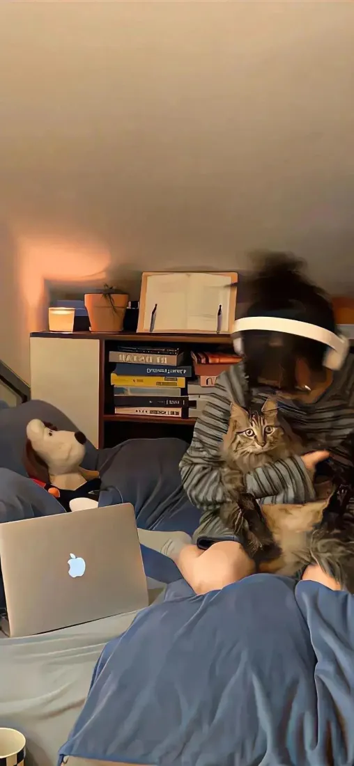 暖光居家室内，戴VR头显的人抱着狸花猫，旁有苹果笔记本与书架。